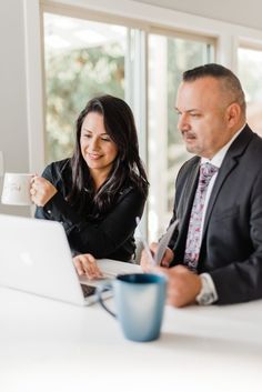 a man and woman sitting at a table looking at a laptop computer, with a cup of coffee in front of them