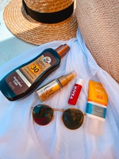 various sunscreens, sunglasses and hat on a white sheet next to a straw hat