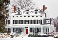 a large white house with black shutters and red door in the middle of winter