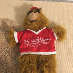 a brown teddy bear wearing a red shirt laying on top of a bed next to a pillow