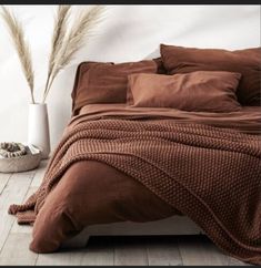 a bed with brown sheets and pillows in a room next to a white vase filled with dry grass