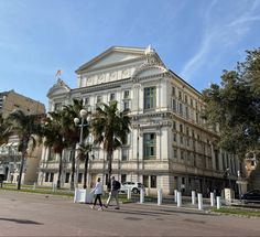 two people walking down the street in front of a large white building with palm trees