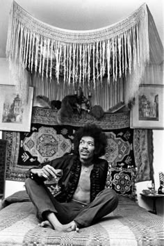 a black and white photo of a man sitting on a bed with fringes hanging from the ceiling