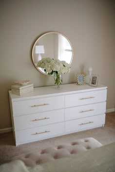 a white dresser with flowers on top and a round mirror above it, in a bedroom