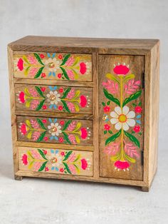 an old wooden cabinet with painted flowers on the doors and drawers, sitting on a white surface