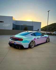 a pink and blue sports car parked in front of a building with the sun setting behind it