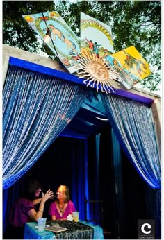 two people sitting at a table in front of a blue canopy with pictures on it