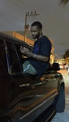 a man sitting on the hood of a car looking at his cell phone while standing next to it