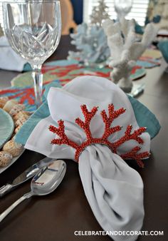 the table is set with silverware, napkins and red coral decorations on it