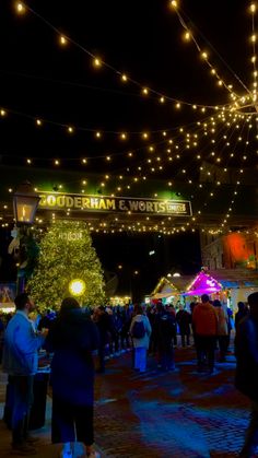 many people are walking around in front of the christmas tree at an outdoor market with lights strung over it