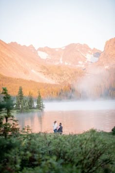 two people are sitting on the shore of a lake with mountains in the background and mist rising from the water