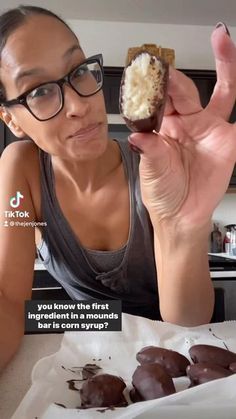 a woman holding up a chocolate covered doughnut in her right hand and looking at the camera