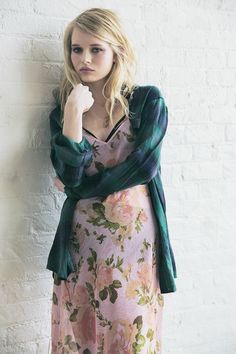 a woman leaning against a white brick wall wearing a floral dress and green cardigan