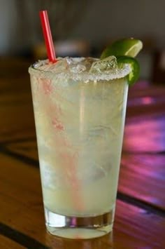 a tall glass filled with ice and lime on top of a wooden table next to a red straw