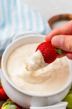 a hand dipping a strawberry into a small white bowl filled with yogurt and strawberries