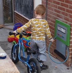 a young boy riding a blue bike next to a building with a water hose attached to the handlebars