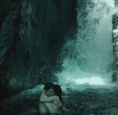 a woman sitting in the water next to a waterfall with her arms around her head