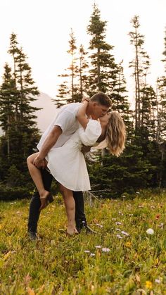 a man and woman kissing in the middle of a field with pine trees behind them