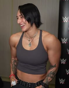 a woman with black hair and tattoos standing in front of a wall wearing a gray tank top