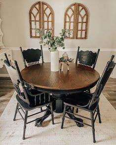 a dining room table with four chairs and a vase on the centerpiece, in front of two arched windows