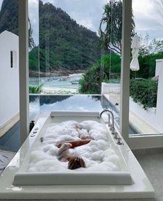 a woman laying in a large bathtub next to a window with mountains behind it