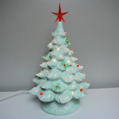 a white ceramic christmas tree with red star on top and lights around the base, in front of a gray background