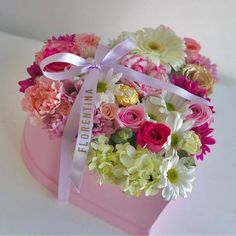 a pink heart shaped box filled with lots of different colored flowers and ribbon tied around it