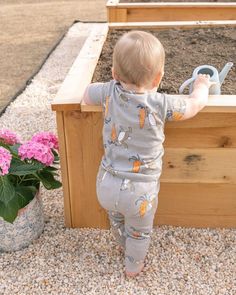 a toddler standing in front of a raised garden bed with flowers and watering can