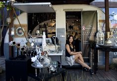 a woman is sitting in the doorway of a small store with many items on display