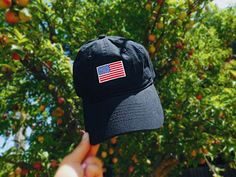 a person holding up a black hat with an american flag sticker on it in front of a tree