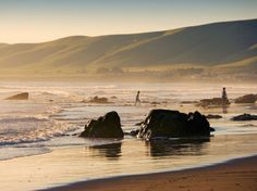 two people are walking on the beach near some rocks and water with mountains in the background