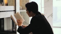 a man sitting down reading a book in front of a fireplace with his hand on the open book