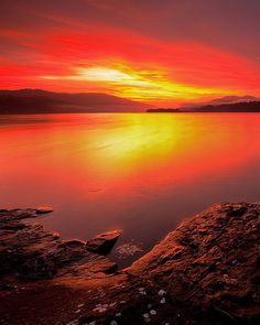 the sun is setting over water with rocks in the foreground and mountains in the distance