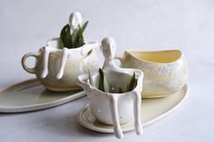 two white vases sitting on top of plates with green plants in the cups and saucers