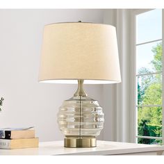 a table lamp sitting on top of a desk next to a book and a window