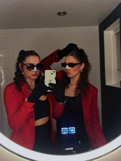 two women wearing sunglasses are looking at their cell phones in front of a bathroom mirror