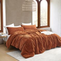 a bed with an orange comforter and pillows in front of two large windows that look out onto the outdoors