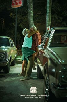 an older man and woman hugging in front of a king king sign at night with cars parked on the street