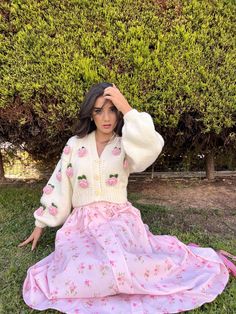 a woman sitting on the ground wearing a pink flowered dress and white cardigan