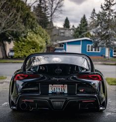 the back end of a black sports car parked in front of a blue house and trees