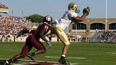 a football player catching a ball in front of an opponent on a field with people watching from the bleachers