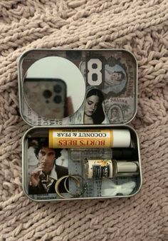 an open tin can with various items in it on a blanket next to a cell phone