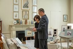 a man and woman standing in a living room looking at pictures on the wall behind them