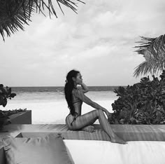 a woman sitting on top of a bed next to the ocean with palm trees in the background