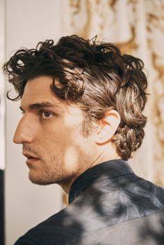 a man with curly hair wearing a black jacket and looking off to the side while standing in front of a wall