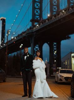 a bride and groom standing in front of the brooklyn bridge at night with their wedding gowns flowing down