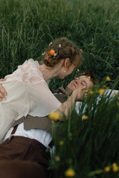 a man and woman laying in the grass with flowers on their head, one is kissing the other's forehead