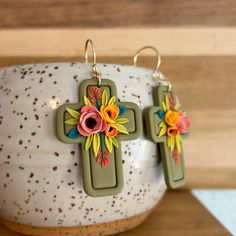 two cross earrings with flowers on them sitting on top of a white vase next to a wooden table