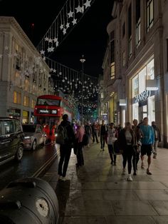 many people are walking down the street in front of buildings at night time with lights strung above them