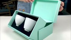 two white cups in a green box sitting on a table next to a person's hand